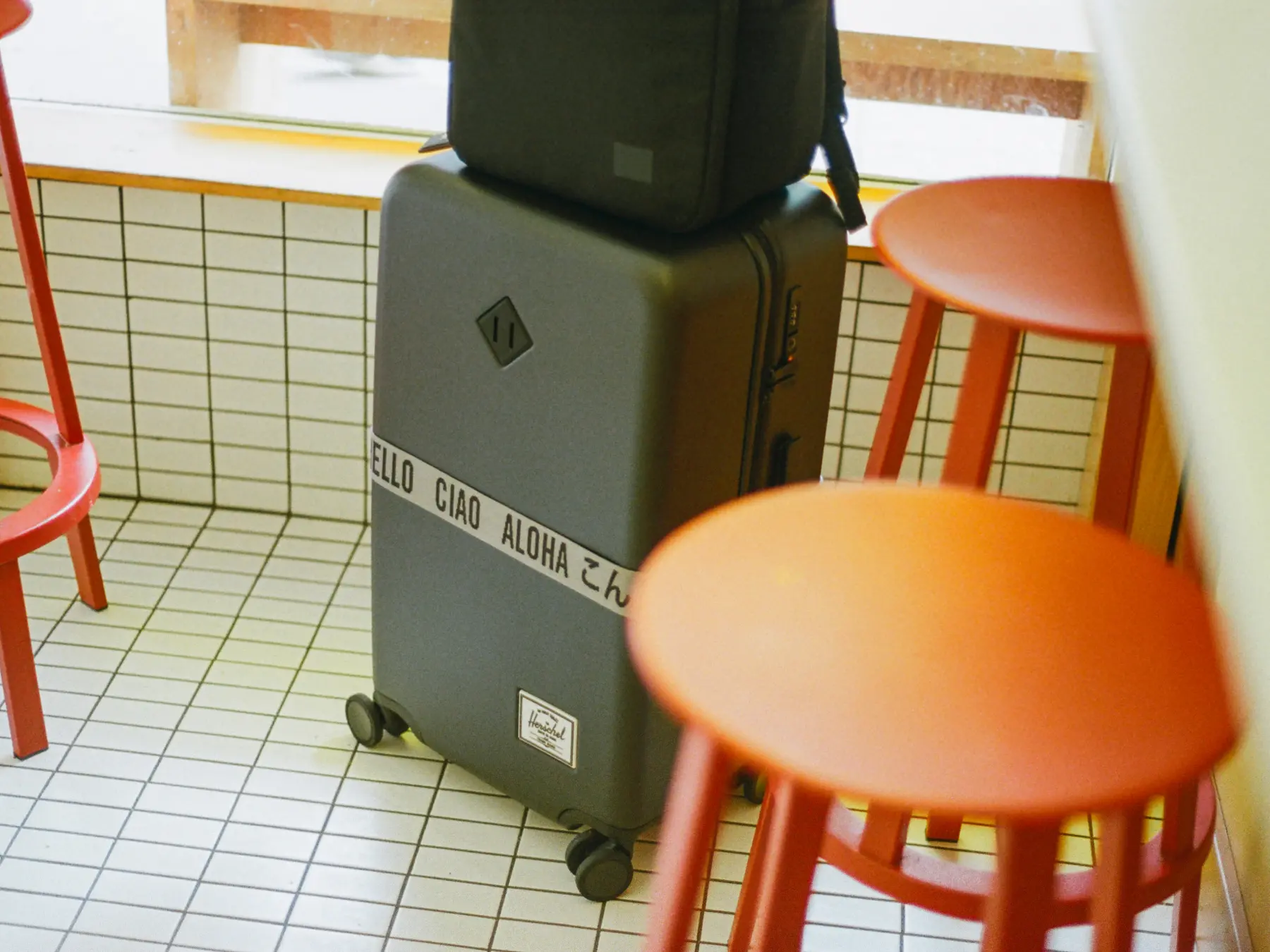 a grey large herschel heritage hardshell luggage with a luggage belt sits in the corner of a cafe 