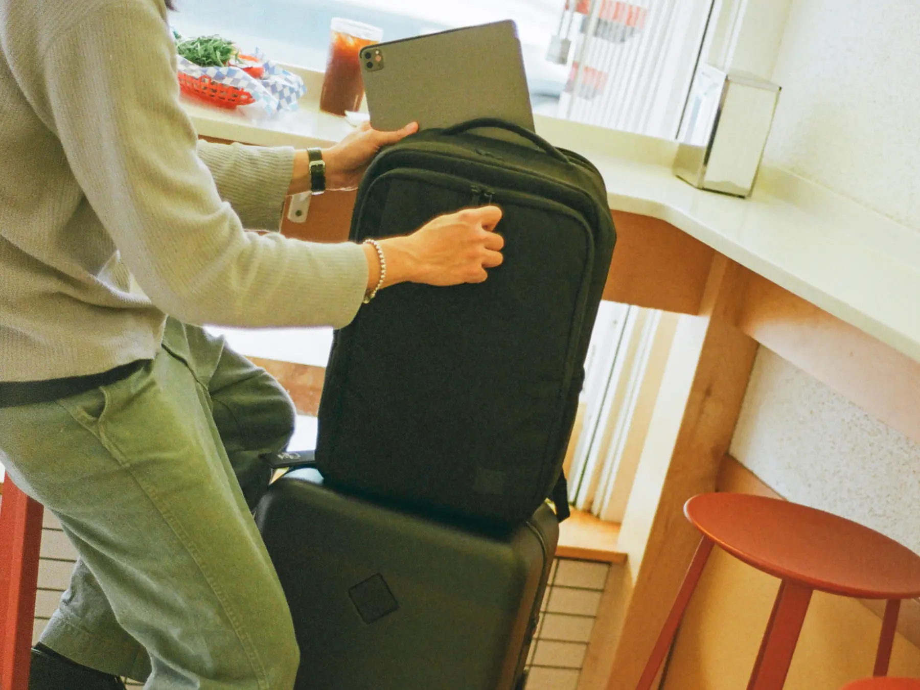 a grey large herschel heritage hardshell luggage with a luggage belt sits in the corner of a cafe. Someone is putting a laptop into a black kaslo daypack that is sitting on top of the luggage.
