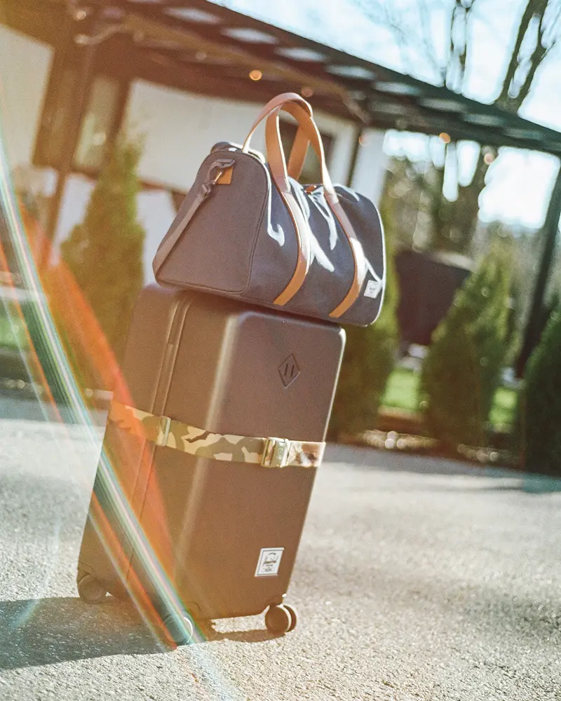 a heritage hardshell suitcase with a novel duffle on it sits in front of a building